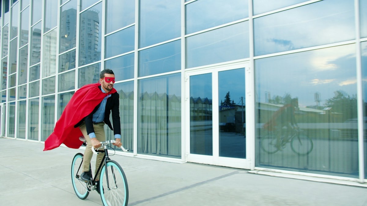 Slow motion of superhero businessman riding bike in business district in city street wearing red cape and mask. Eco transport and work achievements concept.